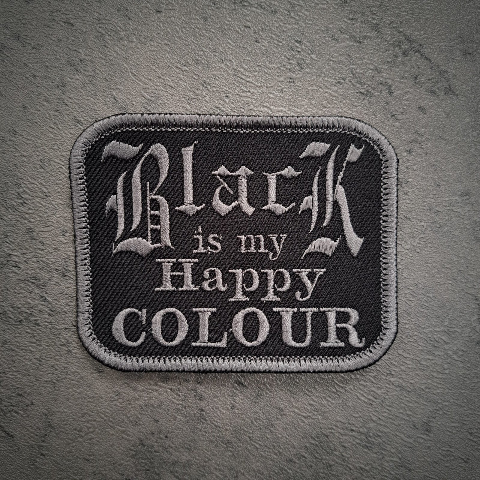 Close-up macro view of an embroidered patch showing the detailed silver stitching of the words "Black is my Happy COLOUR" on black twill fabric.