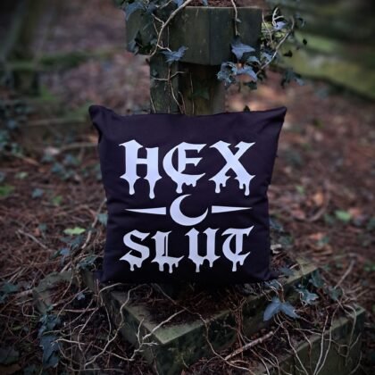 Black gothic cushion cover with “Hex” white dripping gothic lettering resting against an ivy covered stone grave cross in a woodland cemetery setting.