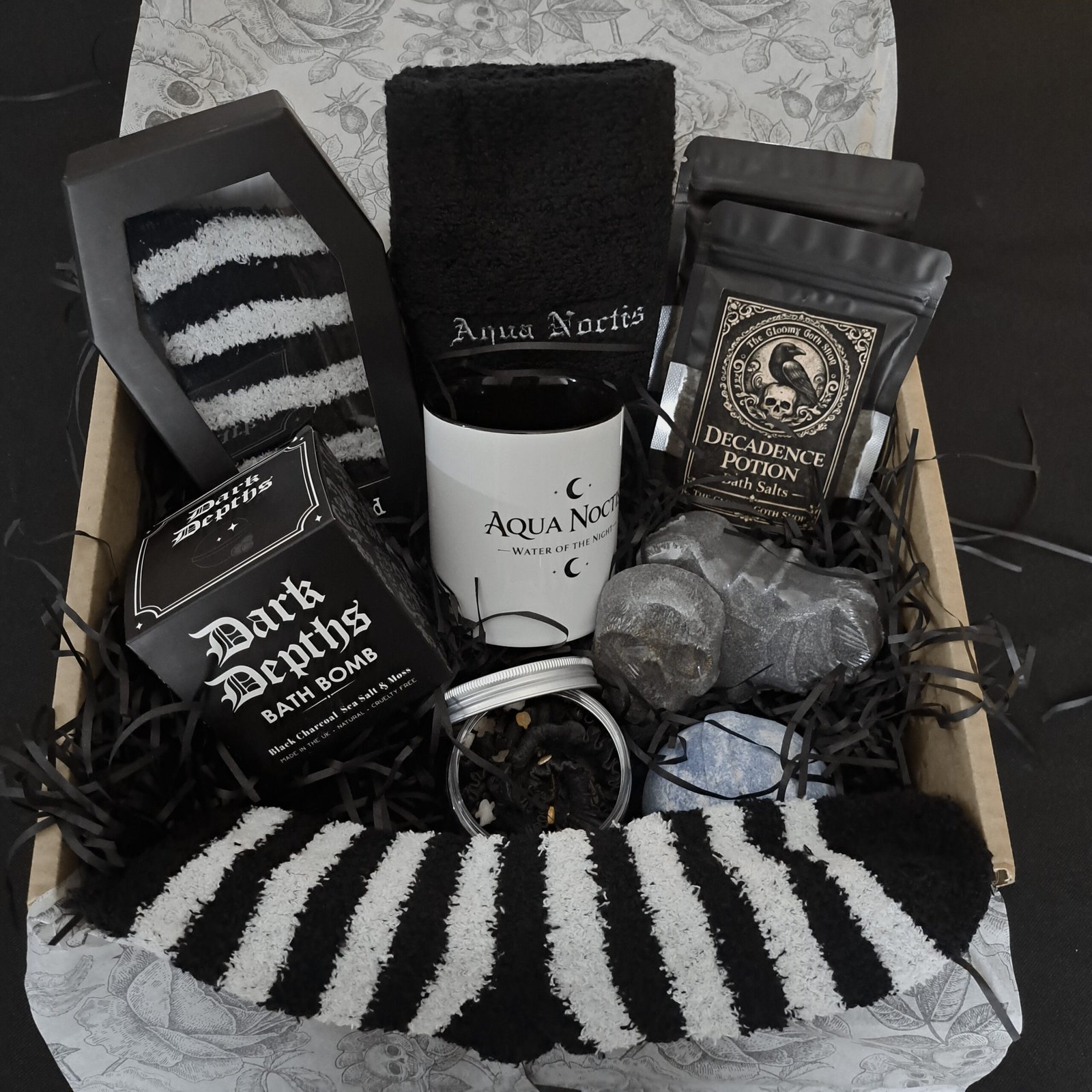 An open 'Aqua Noctis' gothic gift box, packed with spooky self-care items: a coffin-shaped box of fuzzy striped socks, an embroidered black washcloth, a 'Decadence Potion' bath salt pouch, a custom 'Aqua Noctis' mug, and black charcoal and blue bath bombs, all on a bed of black shredded paper.