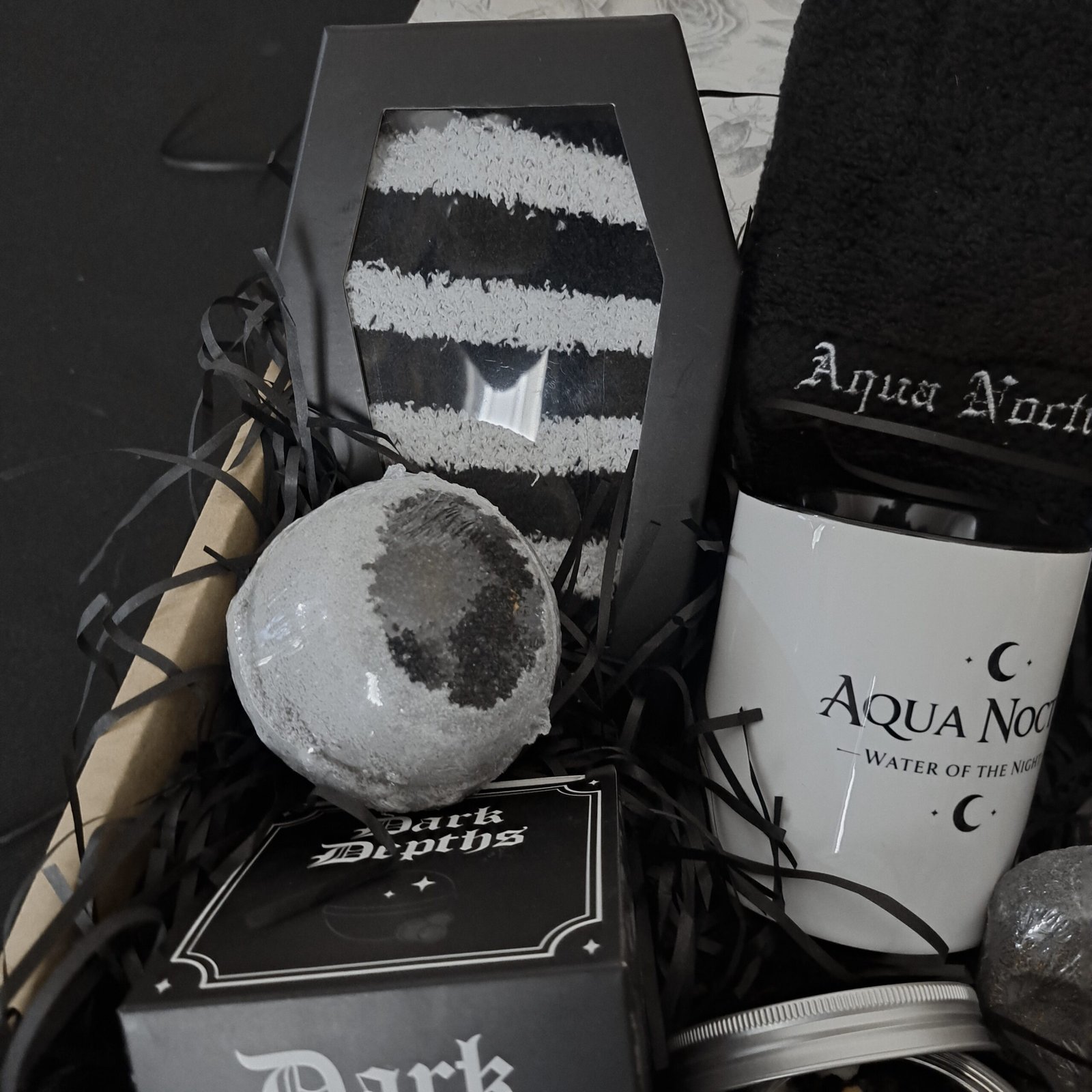 A close-up view of a large, dark charcoal bath bomb, positioned in front of a coffin-shaped box holding fuzzy black and white striped socks. To the side, the white 'Aqua Noctis' ceramic mug is partially visible, all nestled in black packing material.