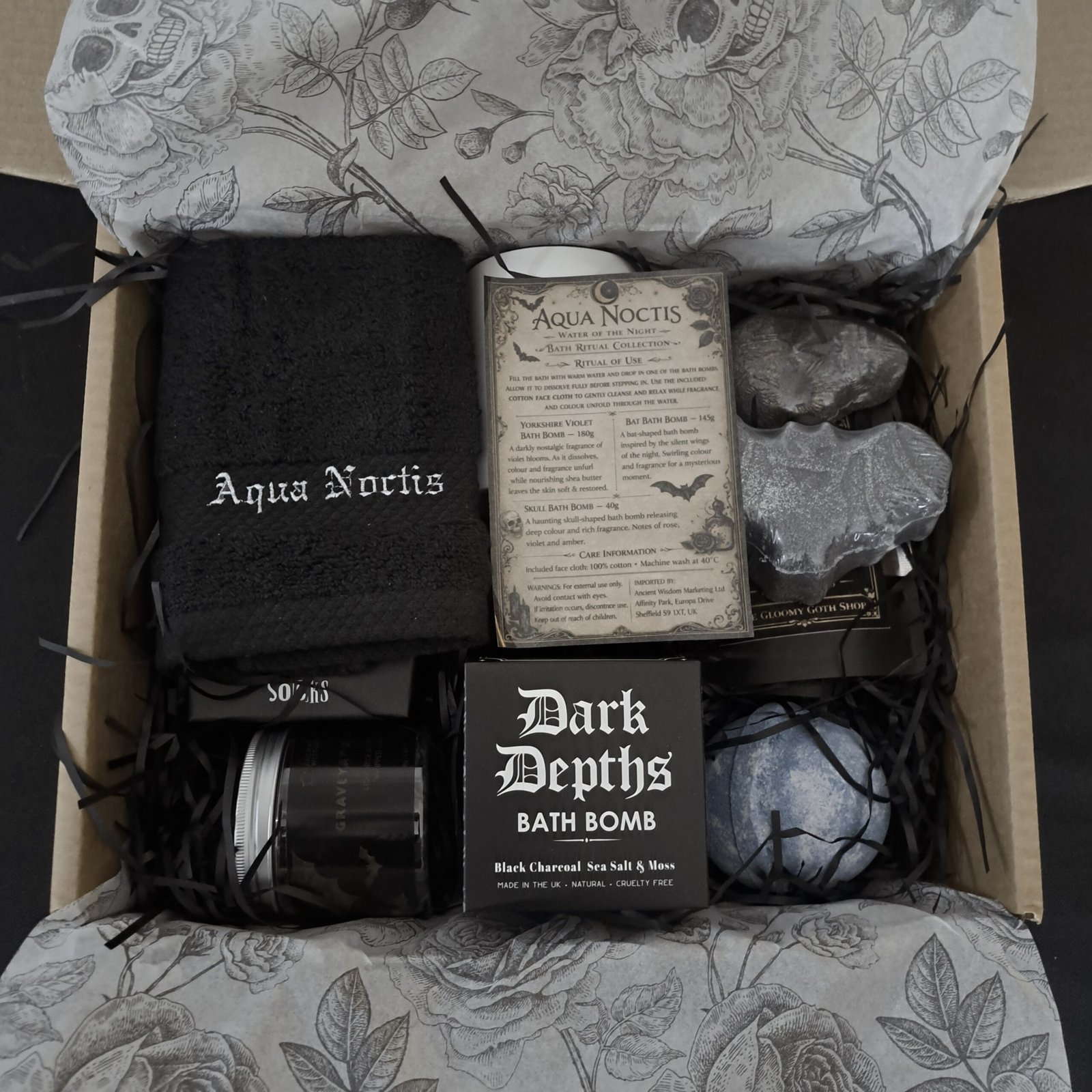 A tidy flat lay of the open 'Aqua Noctis' gift box on a bed of custom black skull-and-rose tissue paper. All items are neatly arranged: the embroidered washcloth, the socks box, bath bombs (skull, bat, blue, charcoal), potion salts pouch, and a clear 'Bath Ritual Collection' care sheet detailing usage instructions.