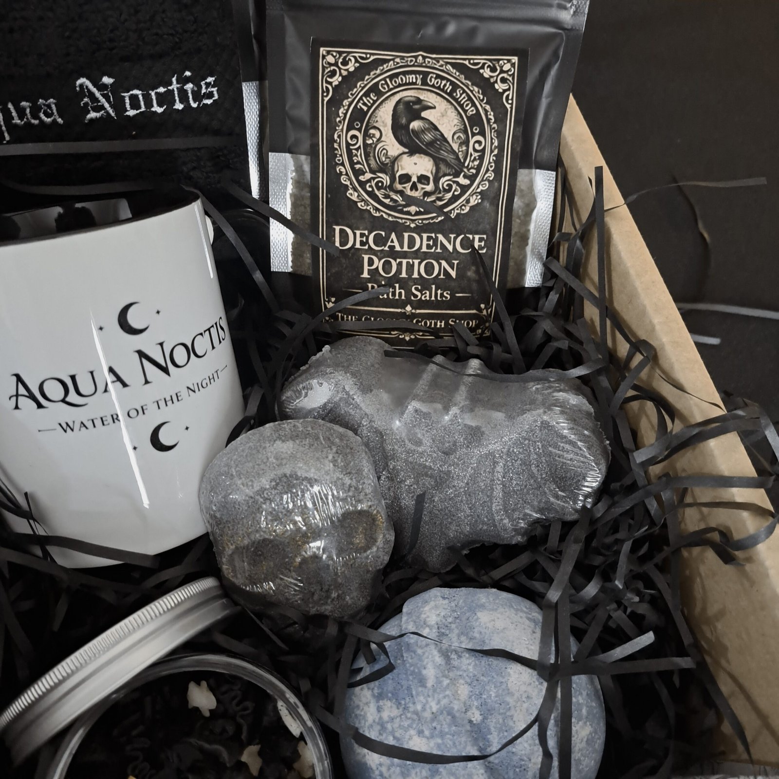 A detailed shot focusing on the 'Decadence Potion' bath salts pouch, featuring elaborate gothic art of a crow and skull. In front are sculpted grey bath bombs in the shapes of a realistic human skull and a bat, alongside a blue speckled spherical bath bomb, all settled on black shredded paper.