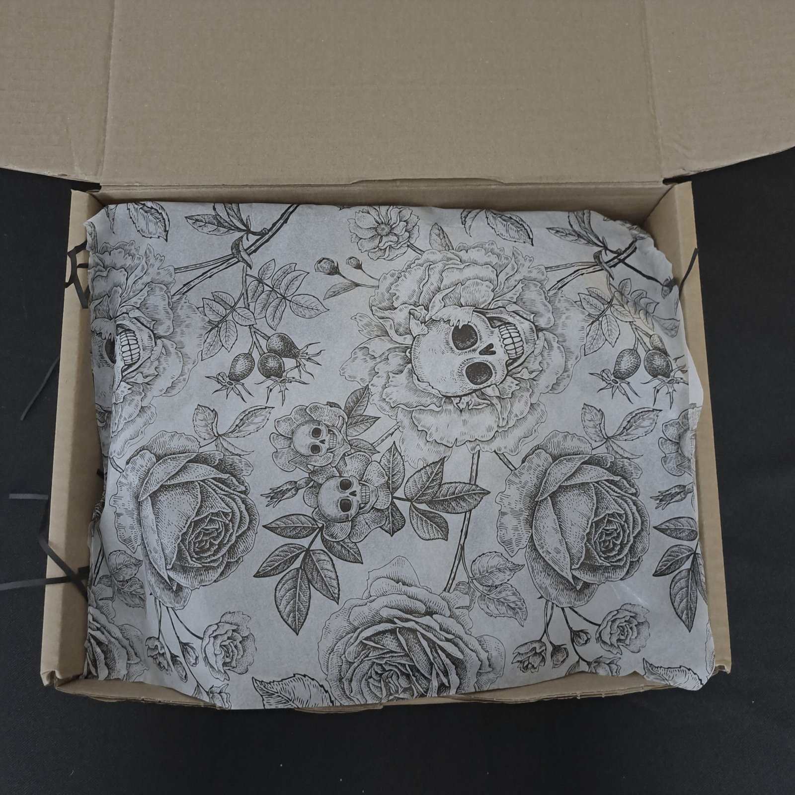 A close-up view looking into an open, cardboard gift box lined with elegant white tissue paper. The tissue paper is printed with a detailed, repeating black pen-and-ink illustration of human skulls nestled among large blooming roses and intricate rose hips.