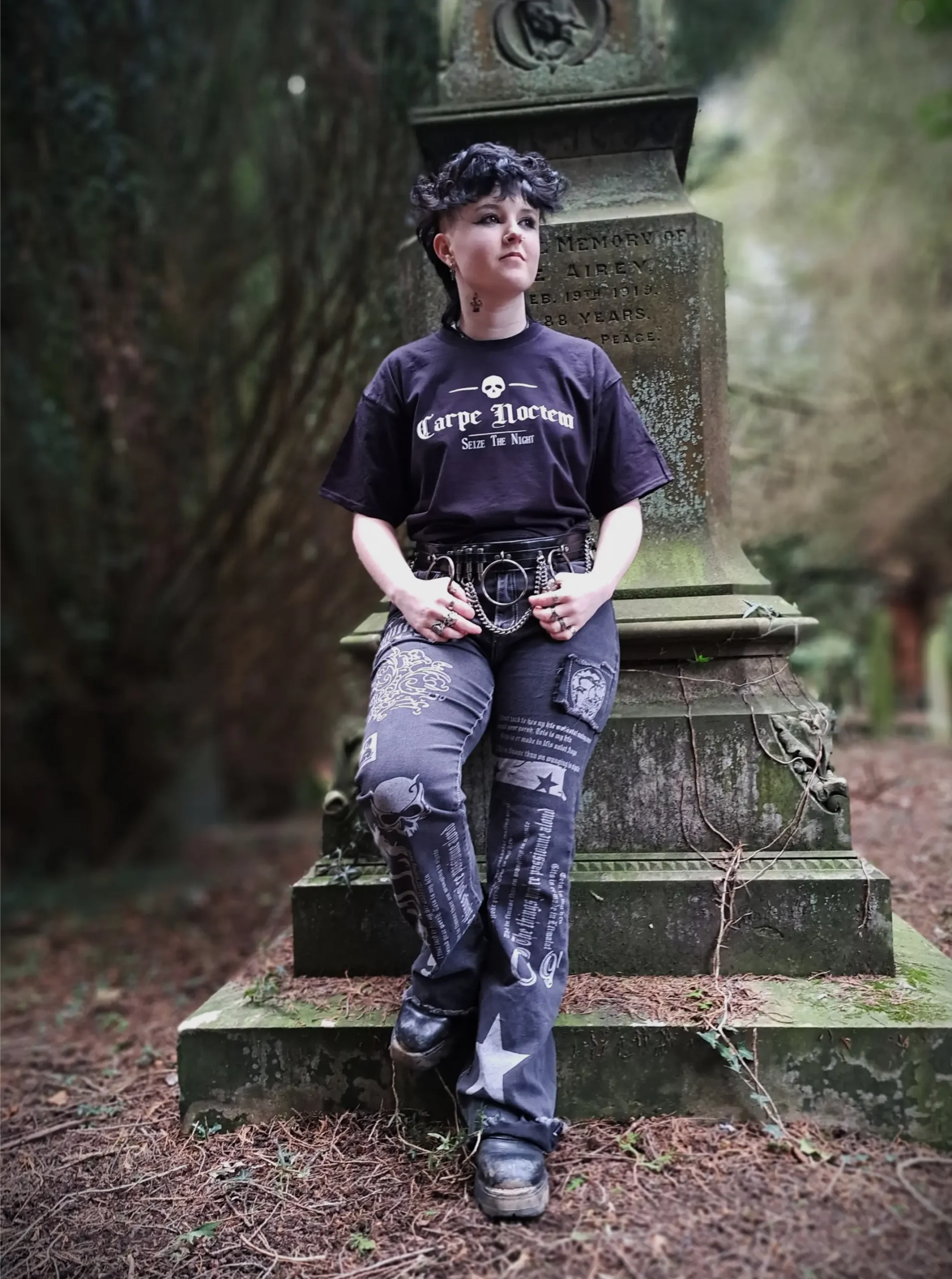Full-length view of a model wearing a black Carpe Noctem T-shirt with patterned gothic trousers, chain belt and boots in a woodland graveyard.