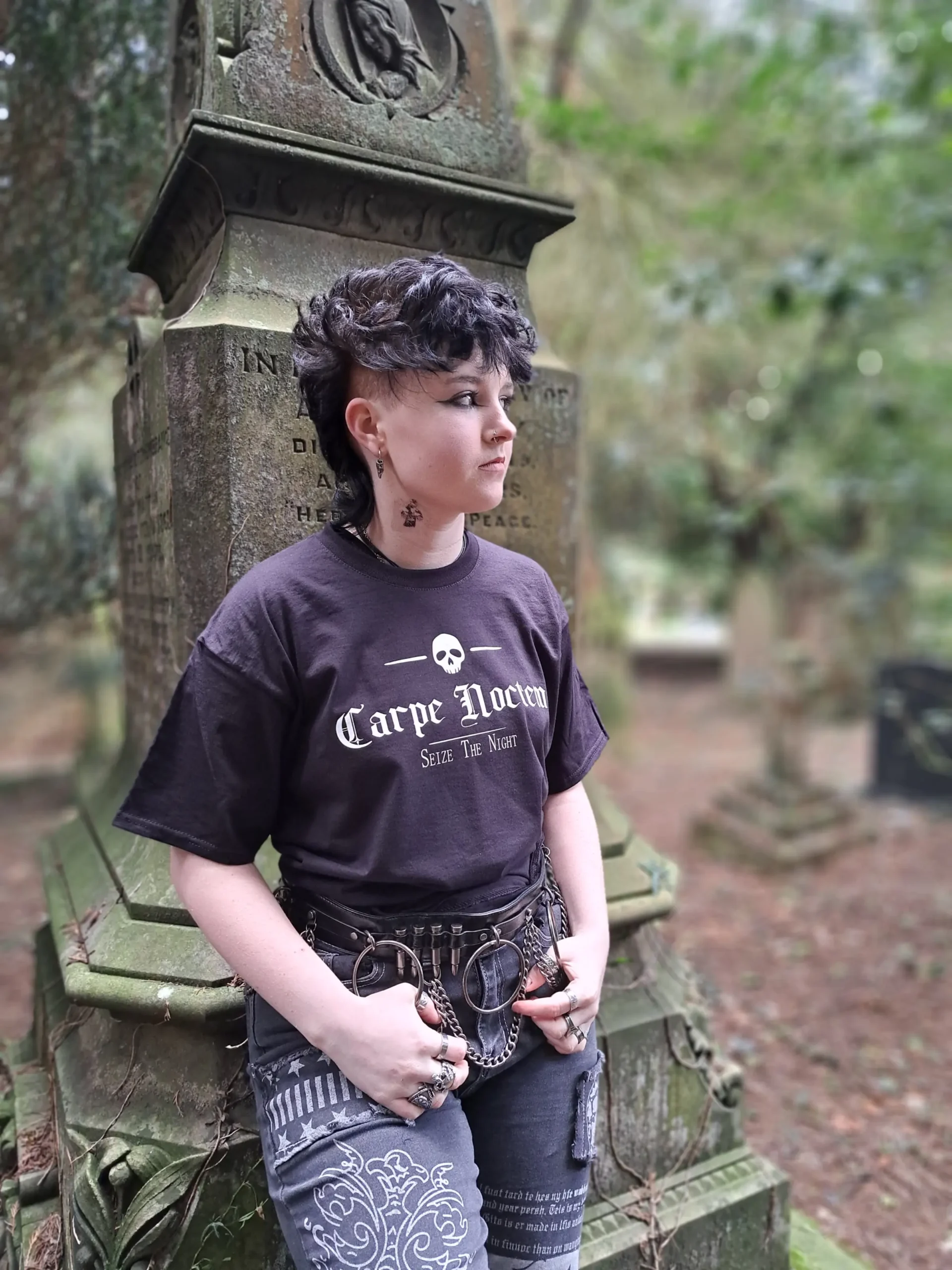 Model wearing a black Carpe Noctem skull T-shirt with gothic lettering, photographed from an angle beside an old gravestone.