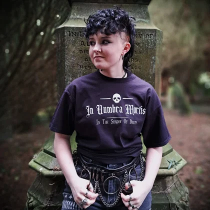 In Umbra Mortis gothic t-shirt in black featuring a white skull and In the Shadow of Death lettering, modelled beside a gravestone.