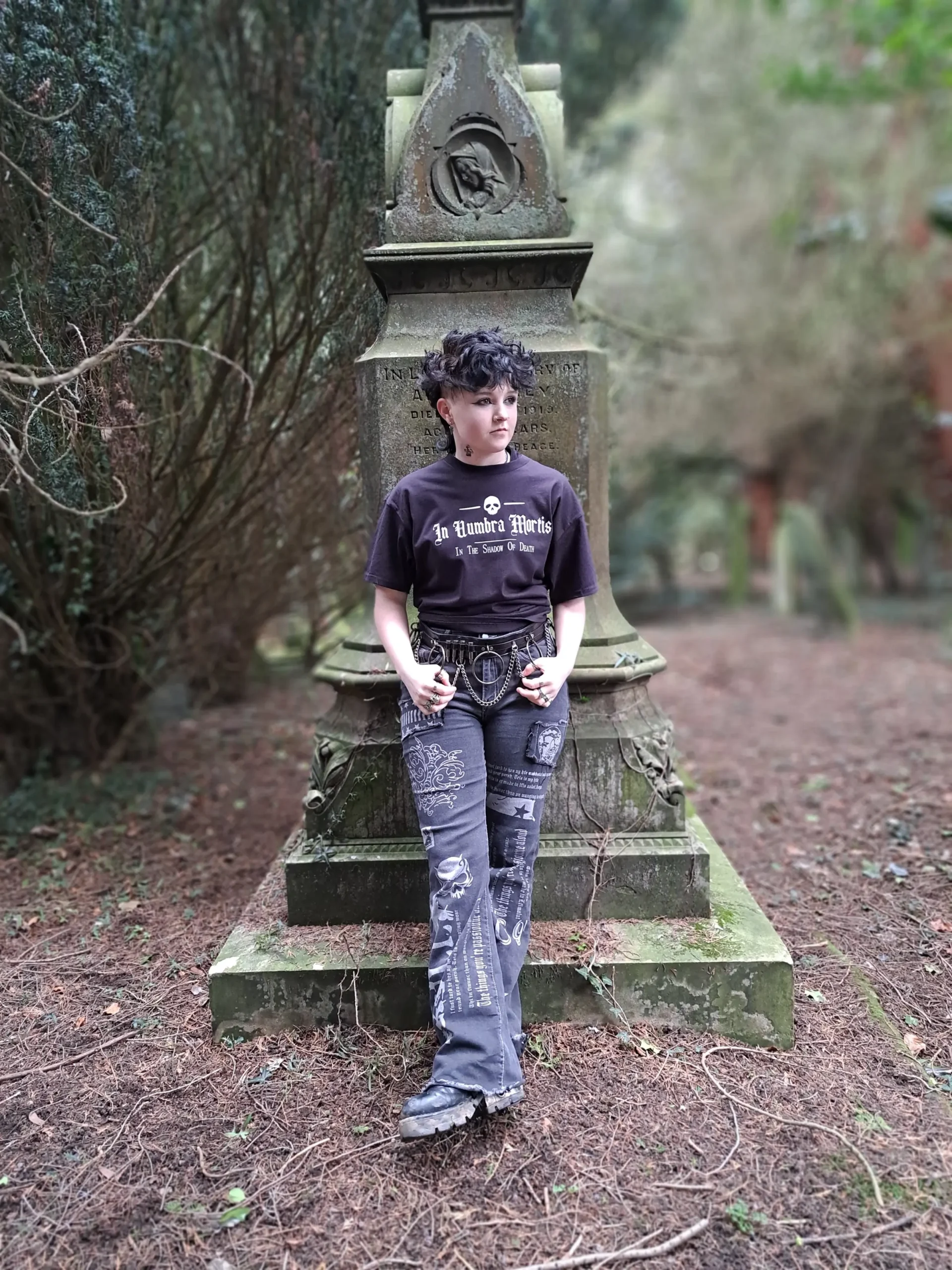 Full-length photograph of a model wearing an In Umbra Mortis gothic T-shirt with dark patterned trousers and boots in front of an old gravestone.