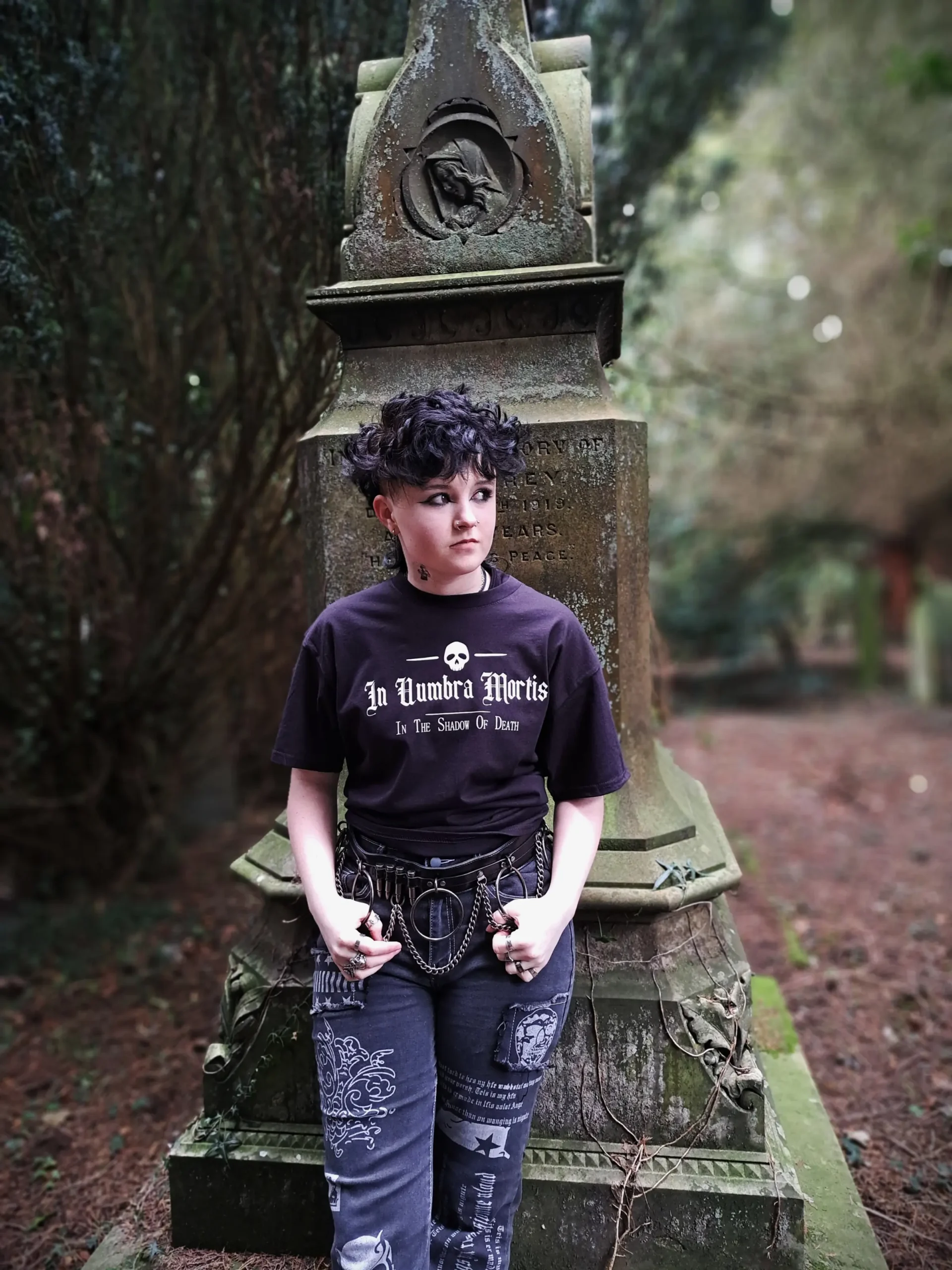 Model wearing a black In Umbra Mortis skull T-shirt with gothic lettering, patterned alternative trousers and chain accessories beside a stone memorial.