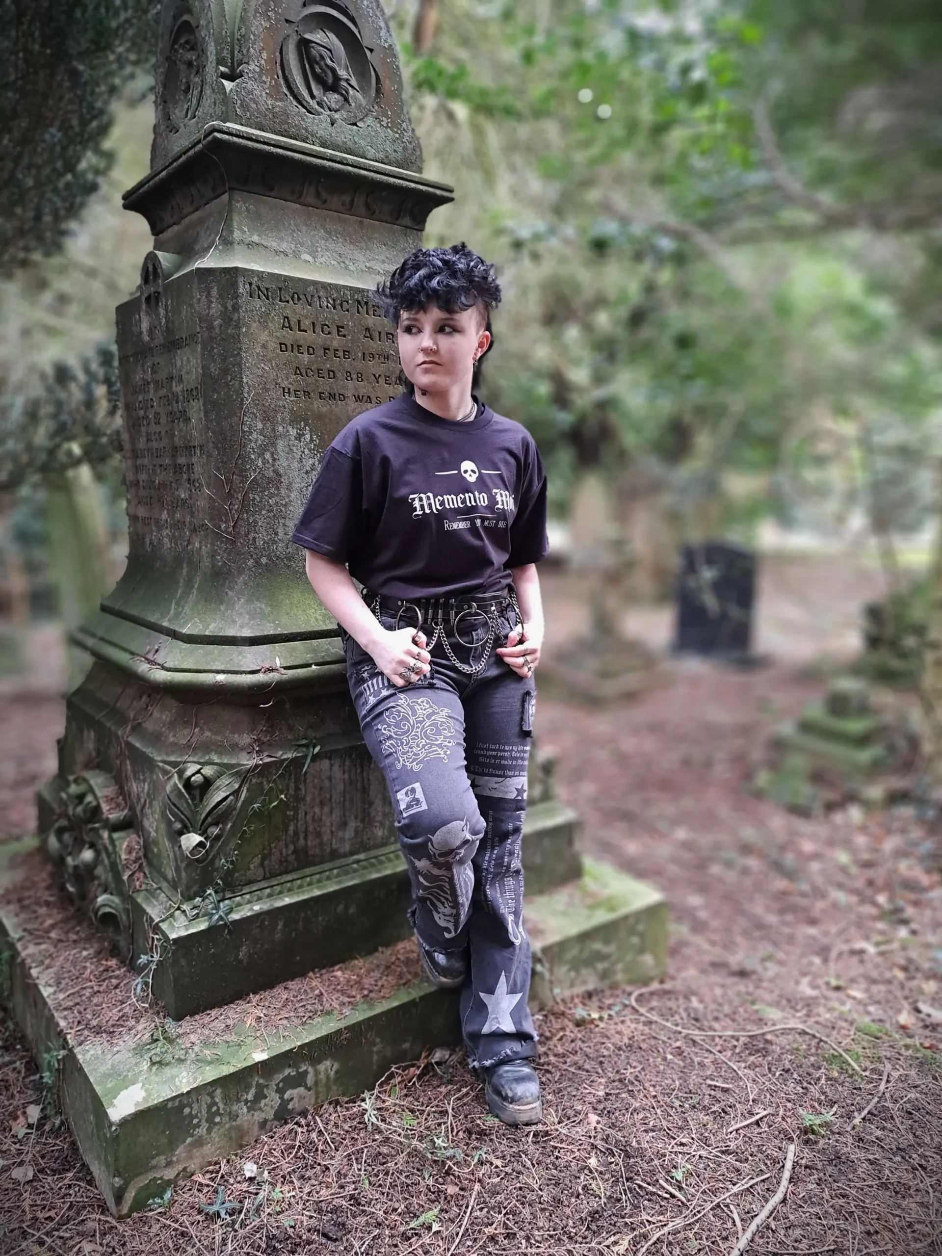 Full-length view of a model wearing a black Memento Mori T-shirt with gothic trousers, chains and boots beside a weathered gravestone.