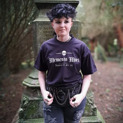 Memento Mori gothic t-shirt in black, featuring a white skull and Remember You Must Die print, modelled beside a gravestone.