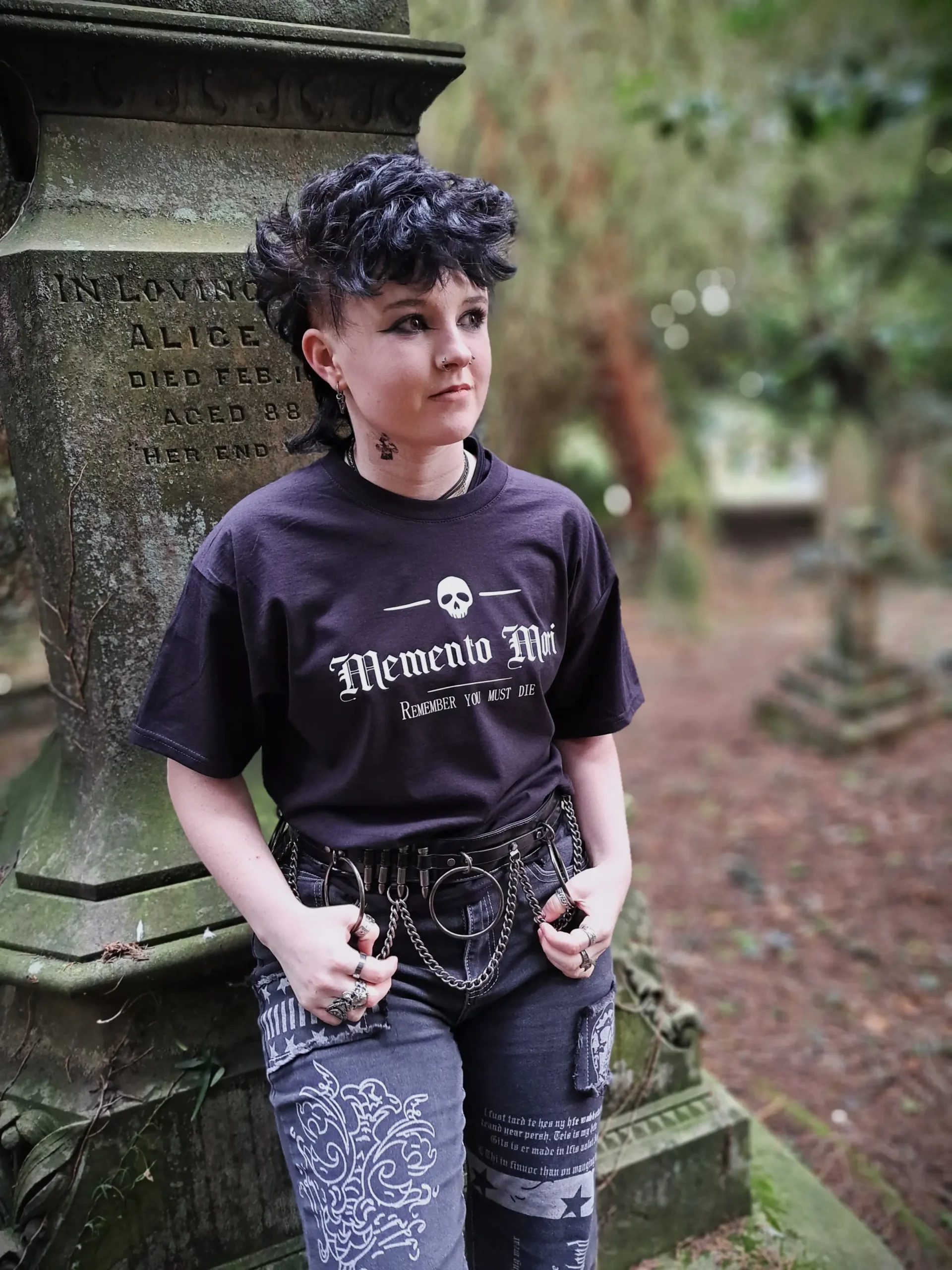 Model wearing a black gothic skull T-shirt with Memento Mori lettering, photographed beside an old stone memorial.